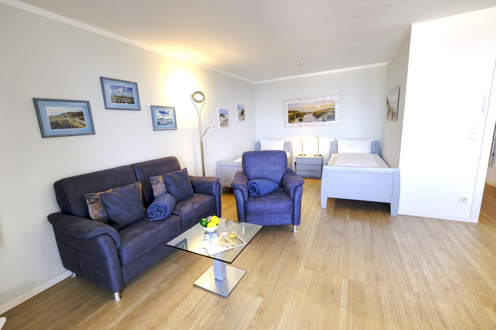 Wohnbereich mit Sofa, Sessel und Bett. Holzboden und Wandbilder.