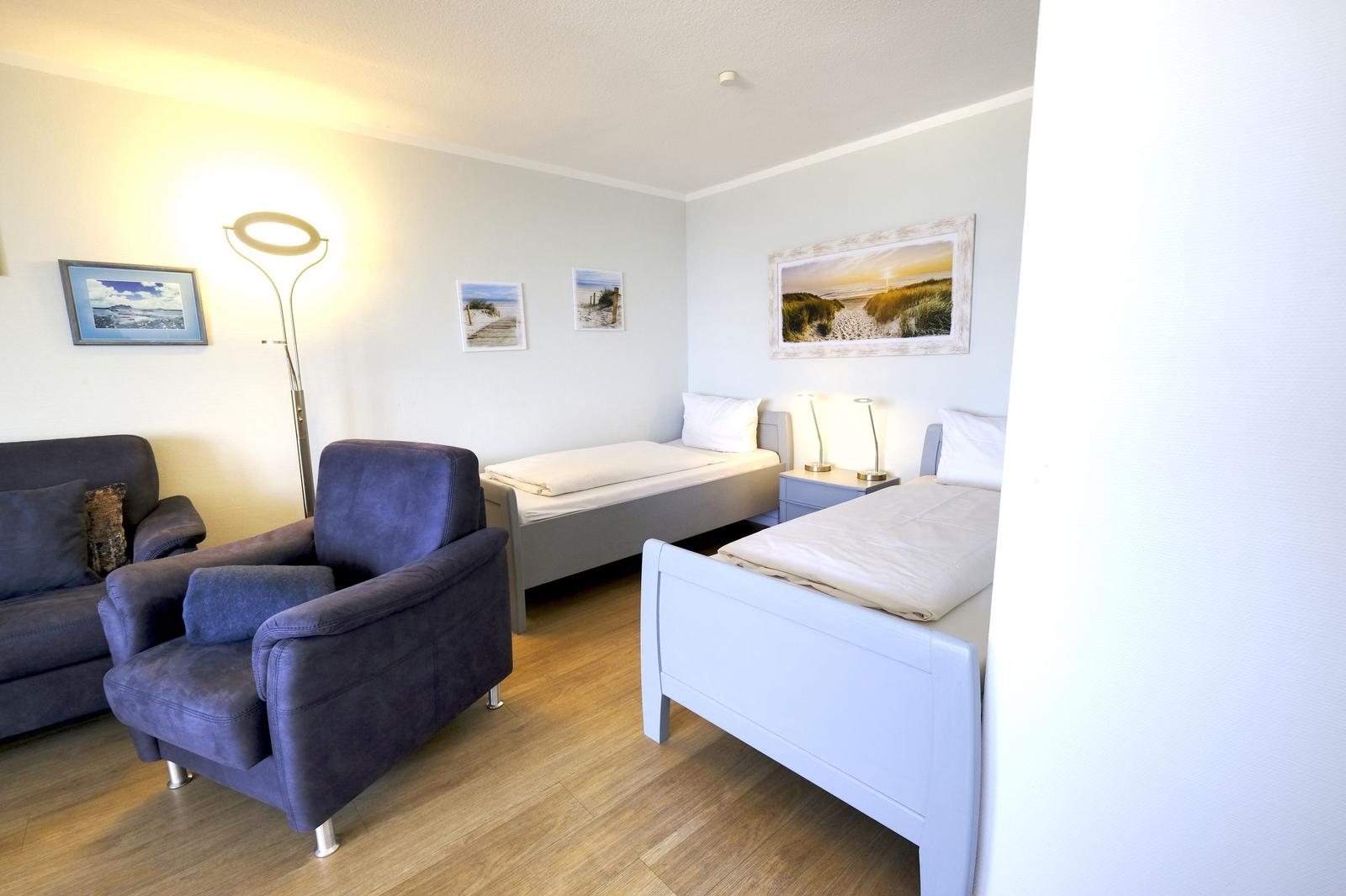 Zwei Betten, Sessel, Stehlampe und Wandbilder in einem Raum mit Holzboden.