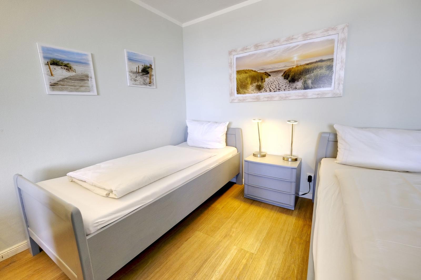 Zwei Betten mit weißen Bettwäsche, Nachttisch mit zwei Lampen, Wandbilder mit Strandmotiven.