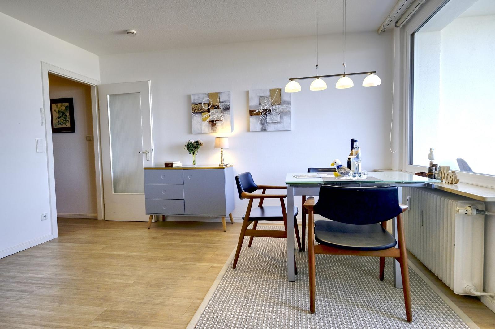 Essbereich mit Tisch, Stühlen und Fenster. Hintergrund: Sideboard und Wandbilder.