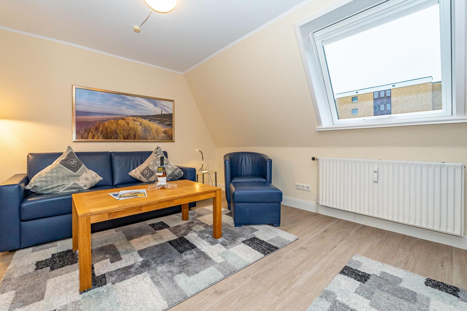 Wohnzimmer mit blauem Sofa, Sessel, Holztisch und Fenster. Wandbild und Teppich.