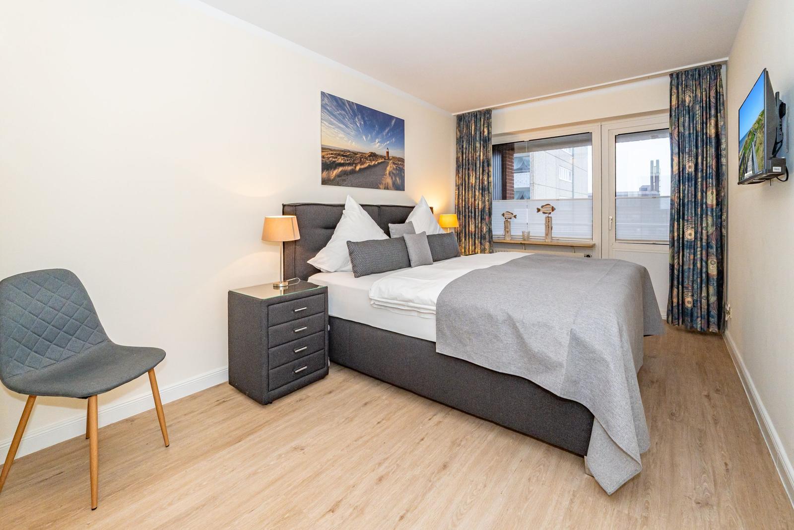 Schlafzimmer mit Bett, Nachttisch, Stuhl und TV. Fenster mit Vorhängen.