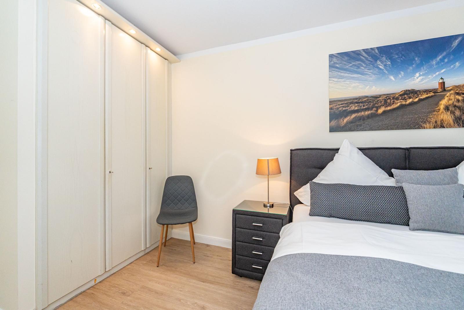 Schlafzimmer mit Bett, Nachttisch, Stuhl und Kleiderschrank. Wandbild zeigt Leuchtturm.