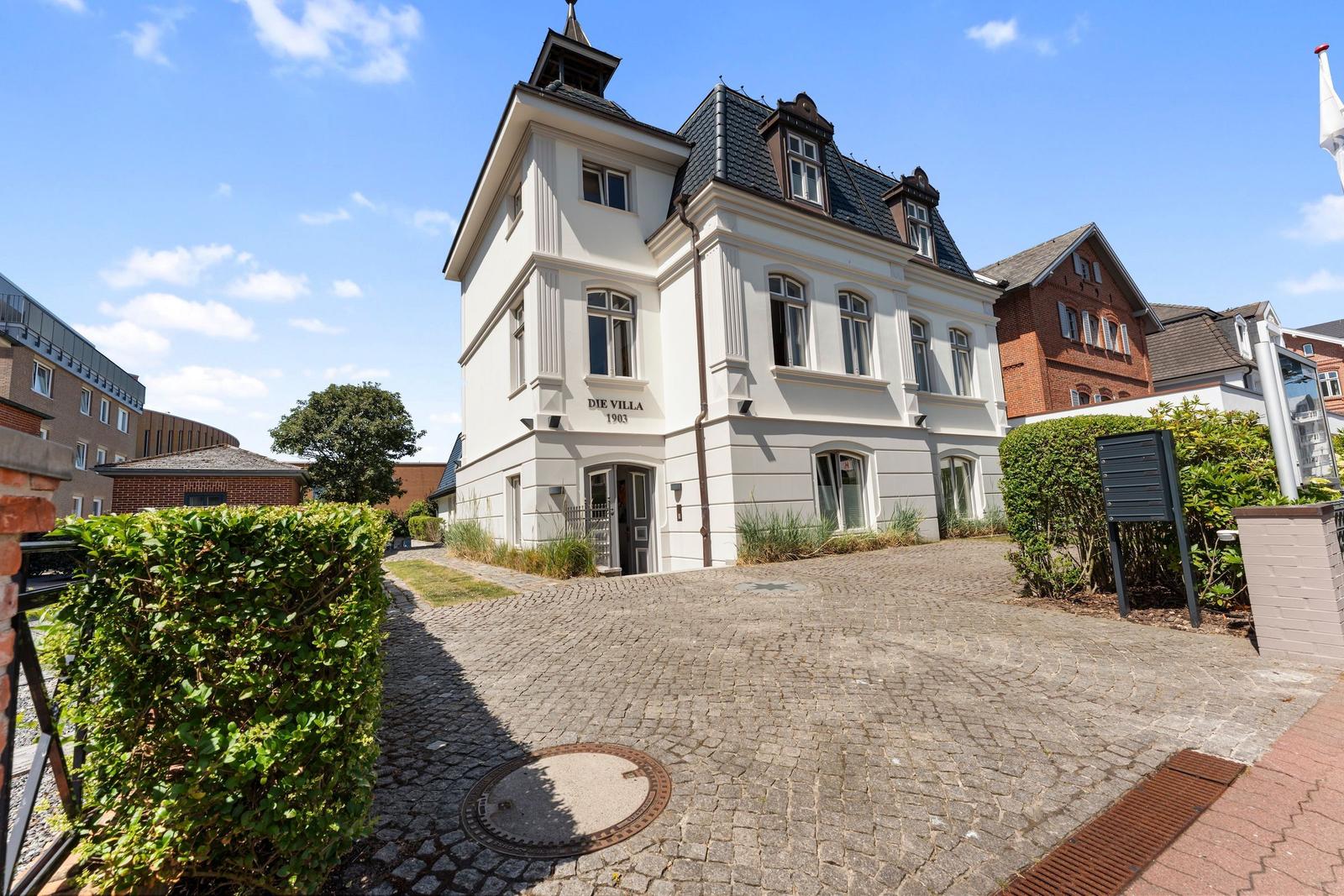 White villa with dark roof and cobblestone driveway. Entrance with sign Die Villa 1903.