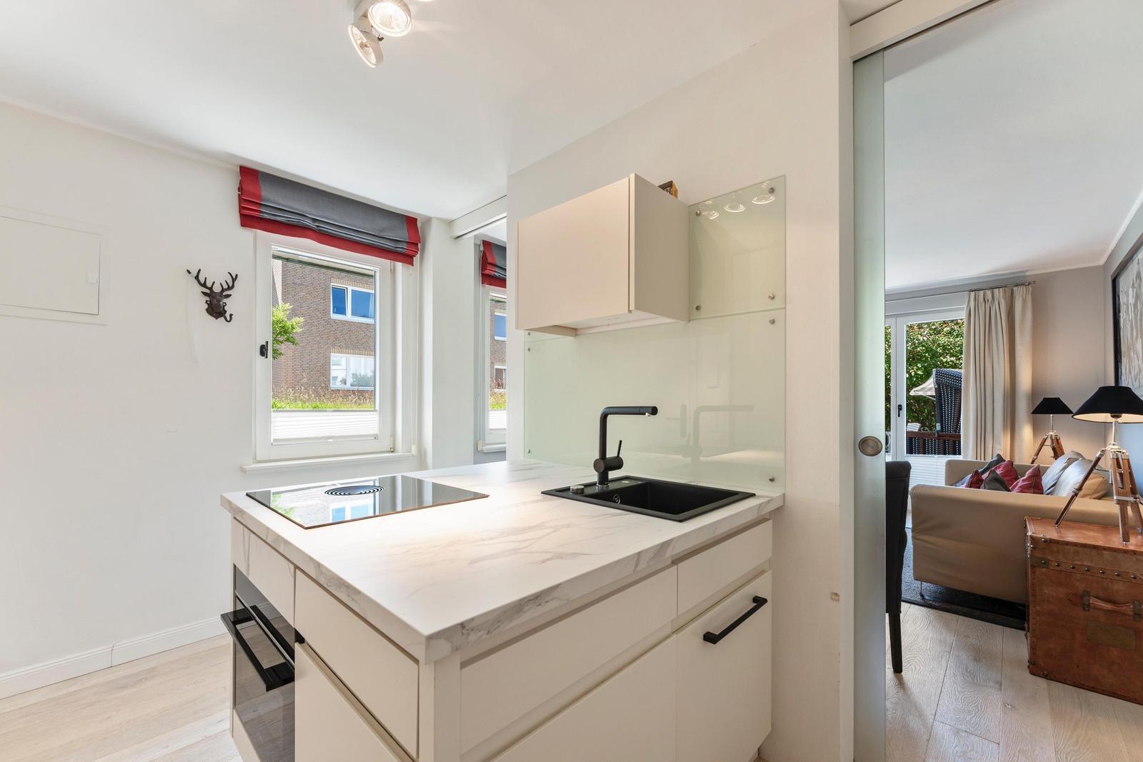 Kitchen with countertop, sink, and window. Passage to living area with sofa and trunk.