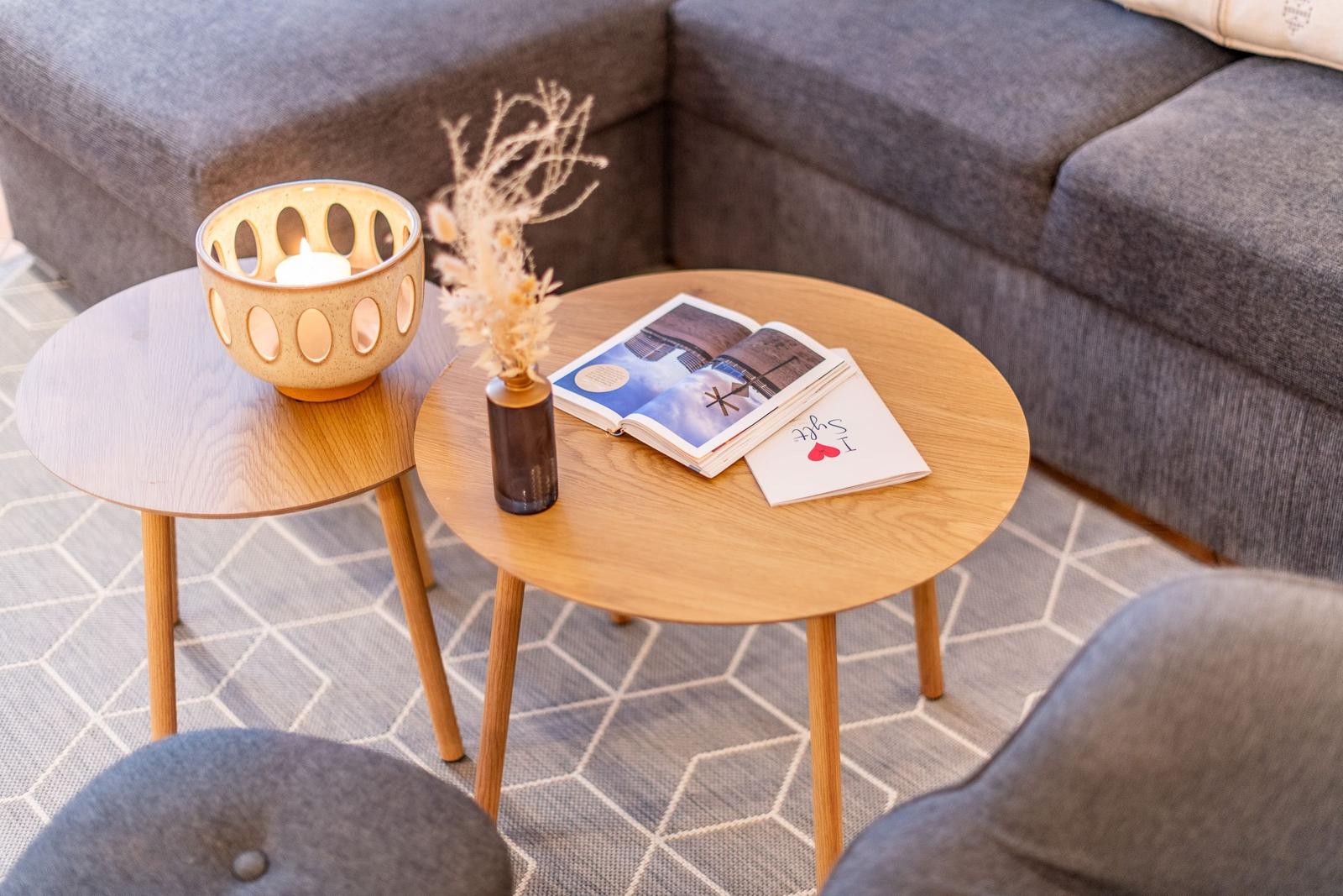 Zwei runde Holztische mit Kerze, Trockenblumen und Buch vor grauem Sofa.