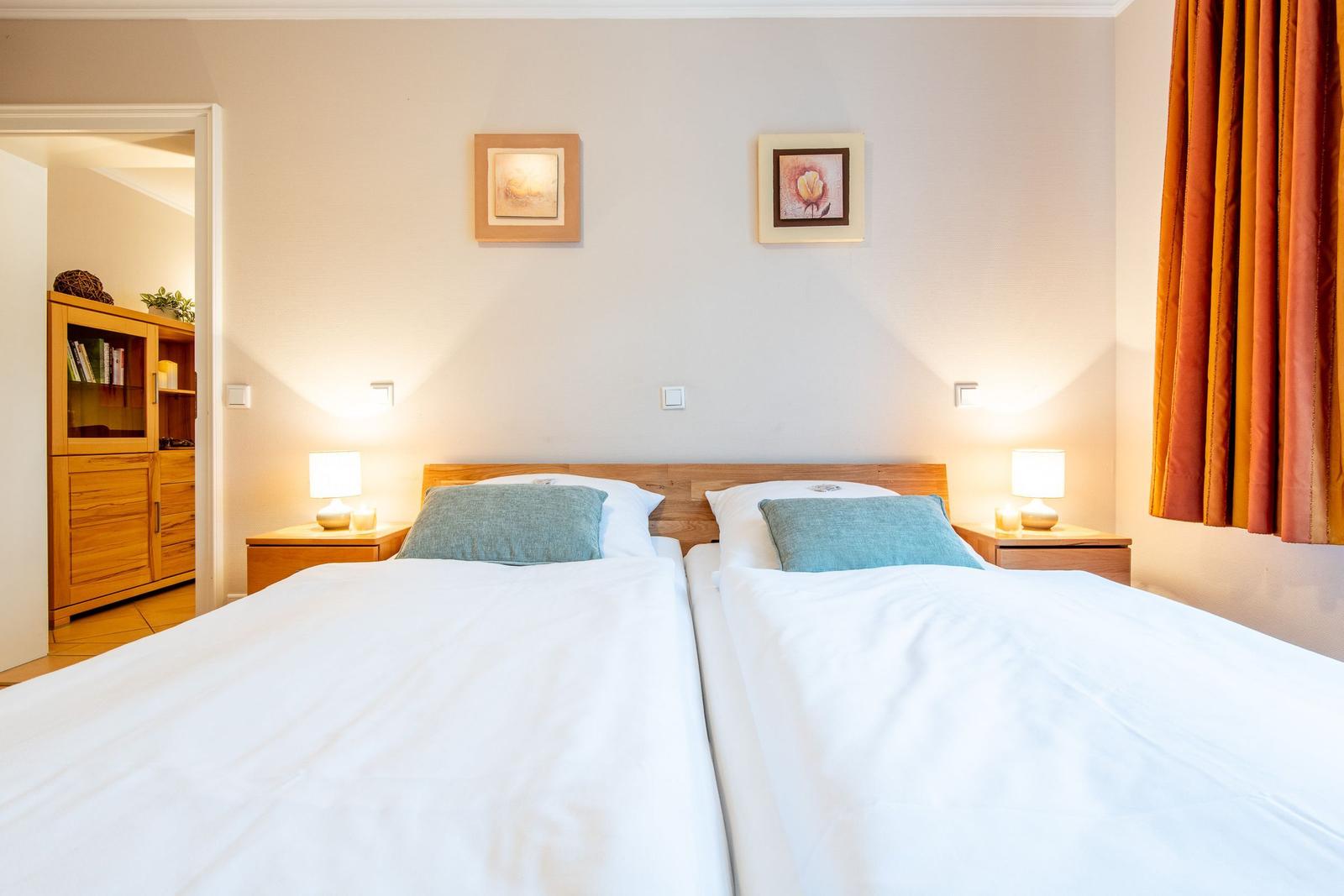 Zwei Betten mit weißen Laken und blauen Kissen. Holz-Nachttische und Bilder an der Wand.
