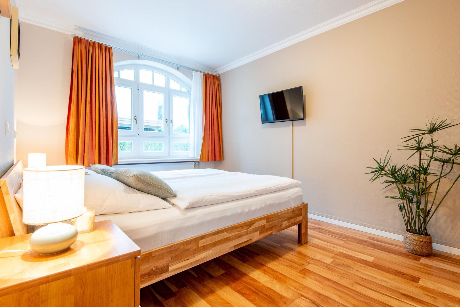 Schlafzimmer mit Holzbett, Fenster, TV und Pflanze.