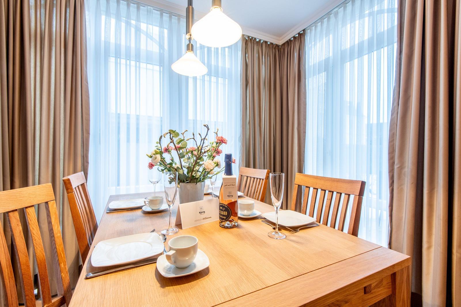 Holztisch mit Stühlen, Blumen und Flasche vor Fenstern mit Vorhängen.