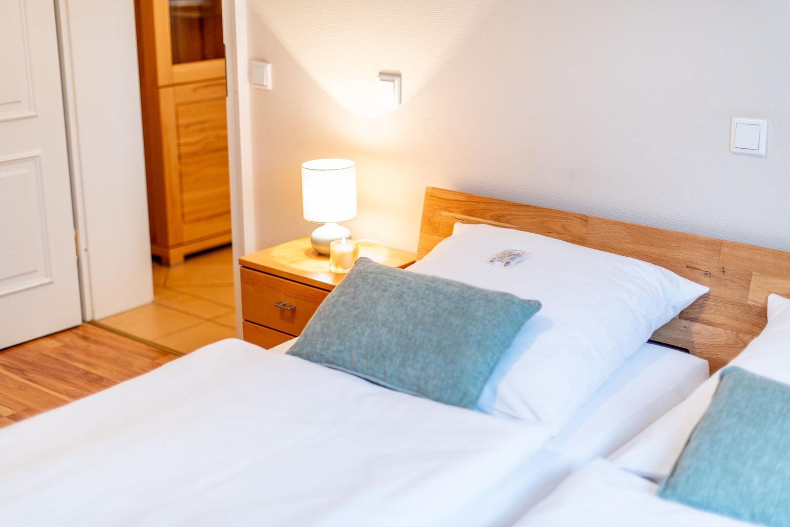 Ein Bett mit weißen Laken und einem grauen Kissen. Neben dem Bett steht ein Nachttisch mit einer Lampe.