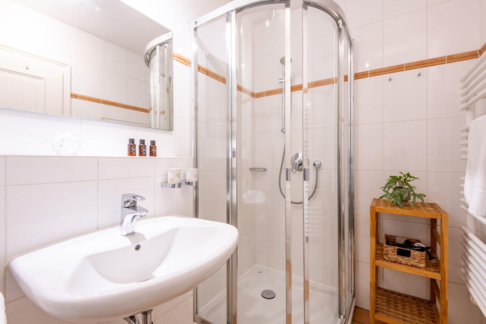 Badezimmer mit Dusche, Waschbecken und Spiegel. Holzregal und Heizung.