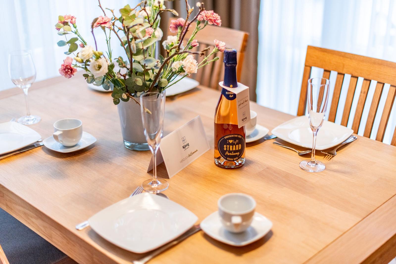 Holztisch mit gedeckten Plätzen, Blumenarrangement und Flasche Sekt.