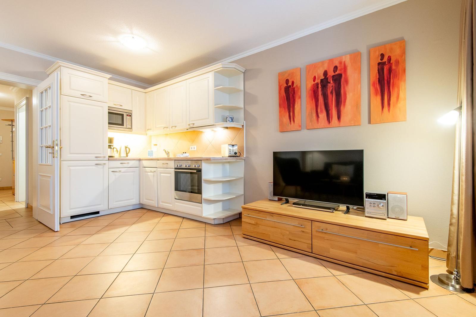 Küche mit weißen Schränken und Wohnbereich mit Fernseher und Wandbildern.