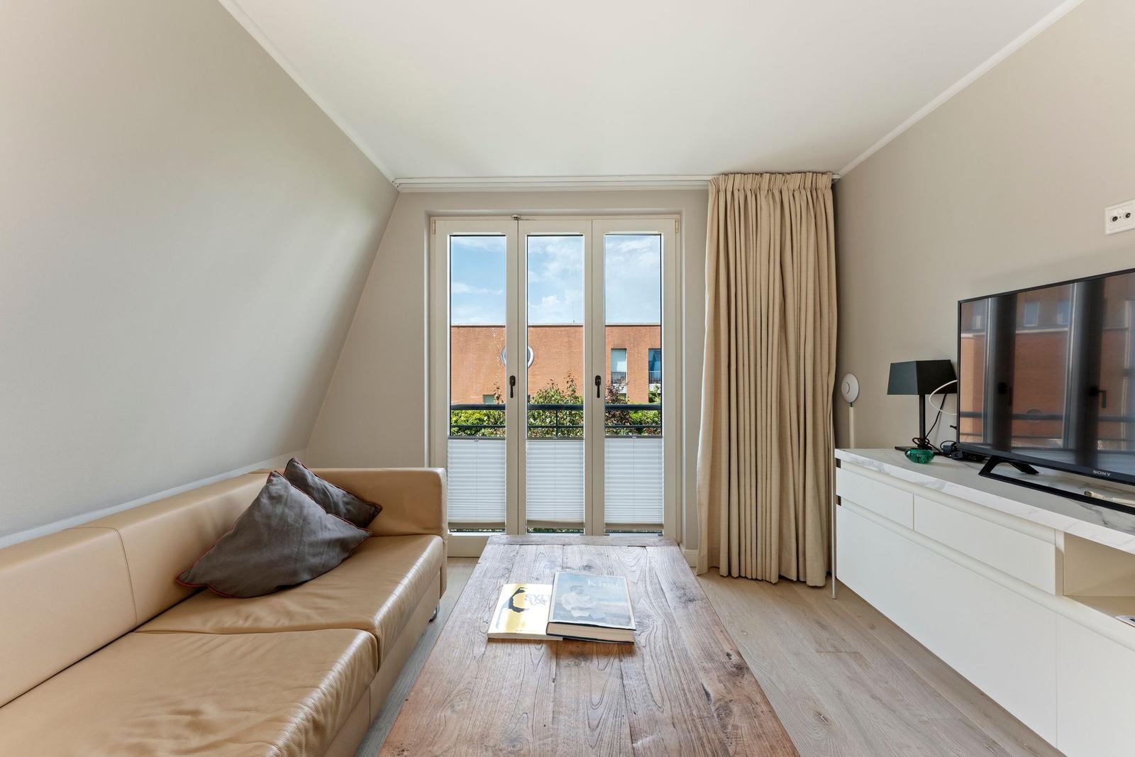 Wohnzimmer mit Sofa, Couchtisch und TV. Balkon mit Blick auf Gebäude.