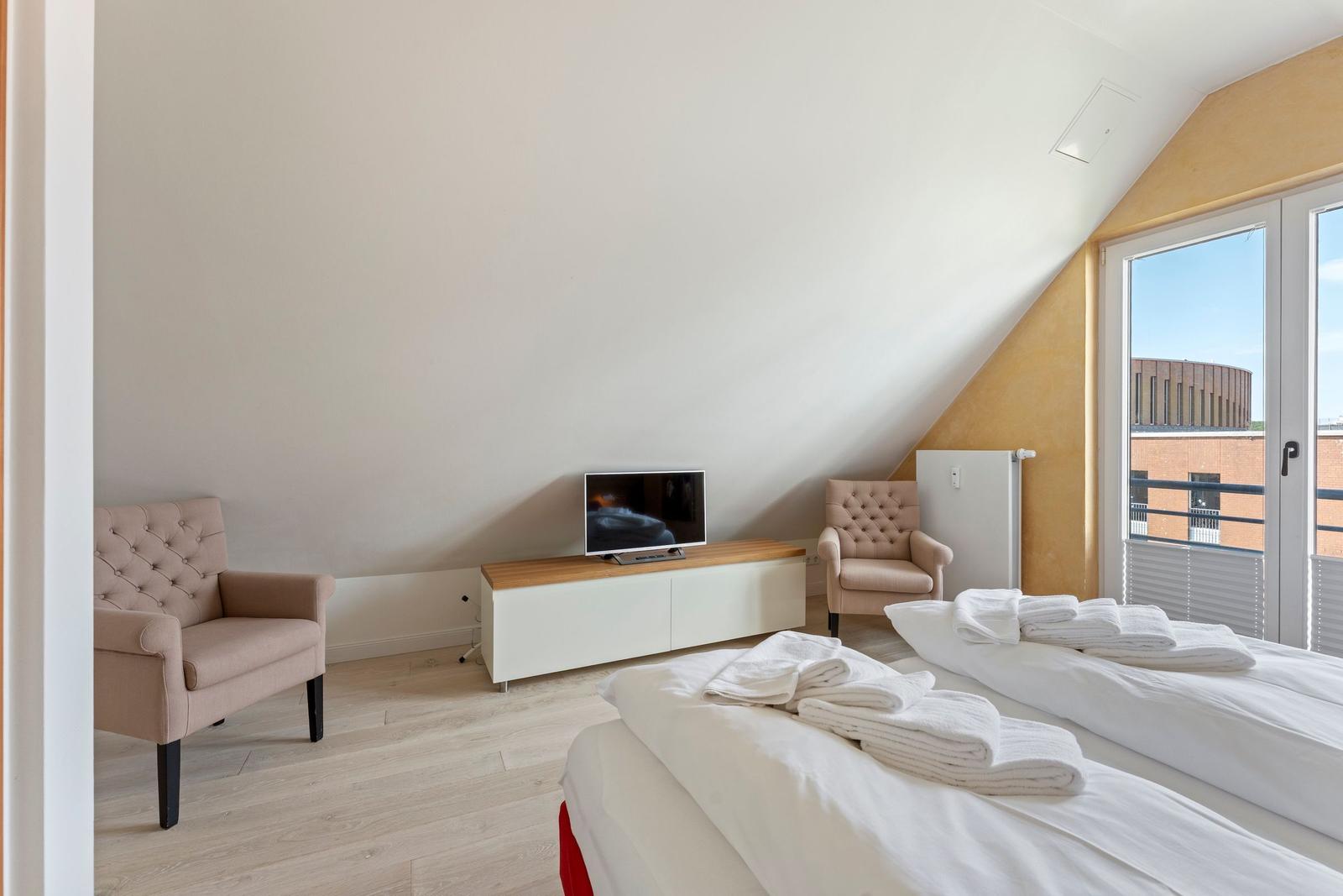 Zimmer mit zwei Betten, TV und Sesseln. Fenster mit Blick auf Gebäude.