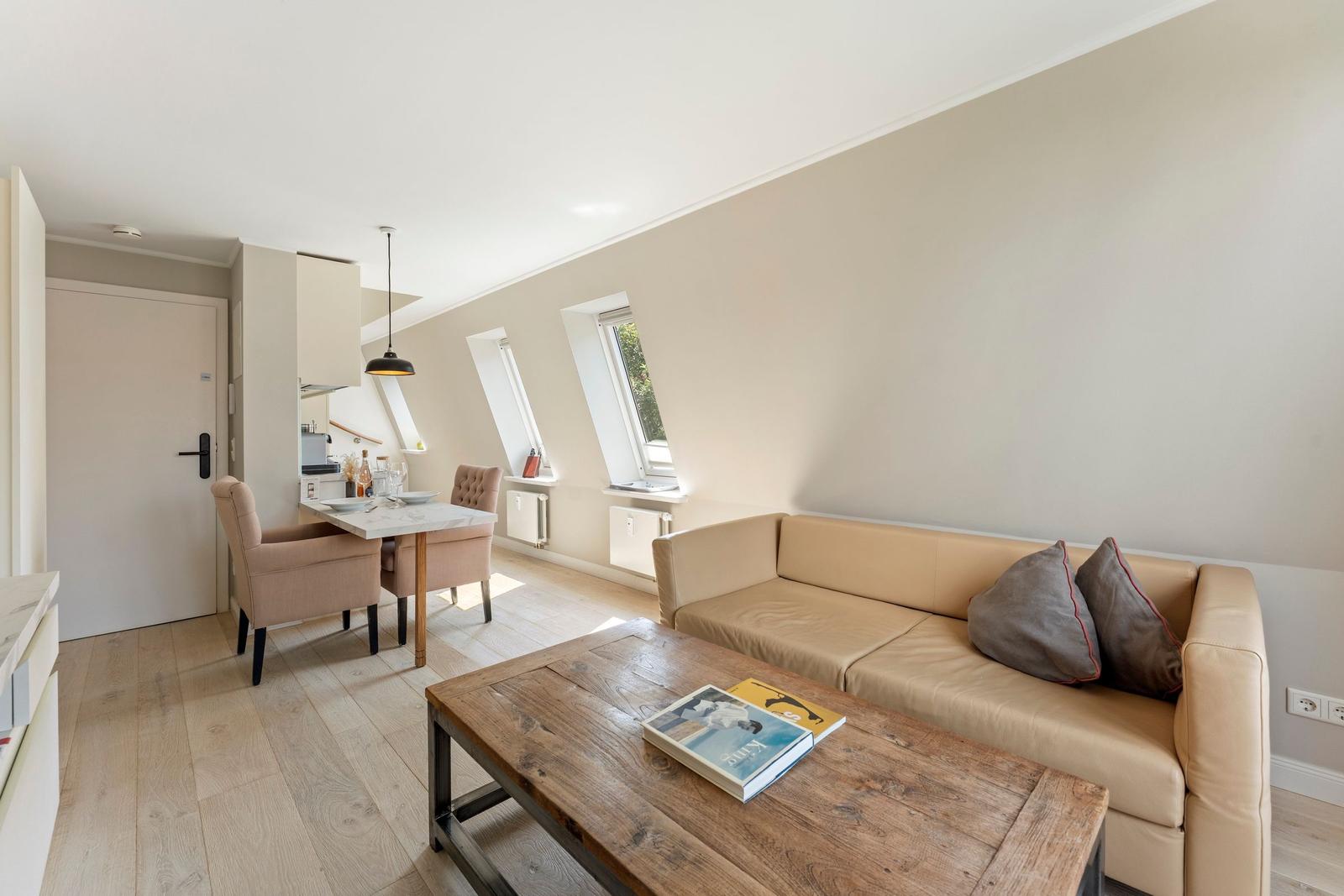 Wohnbereich mit Sofa, Esstisch und Fenstern. Holzboden und helle Wände.