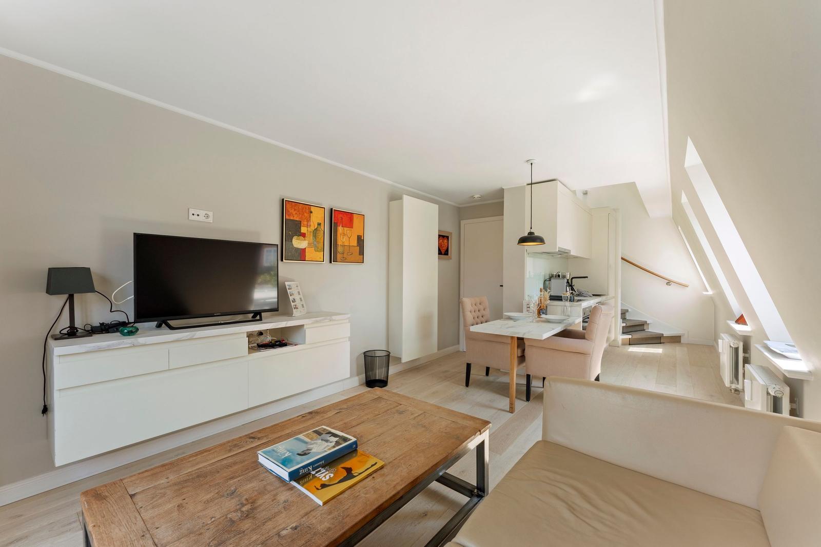 Wohn- und Essbereich mit TV, Couch und Tisch. Küche und Treppe sind sichtbar.