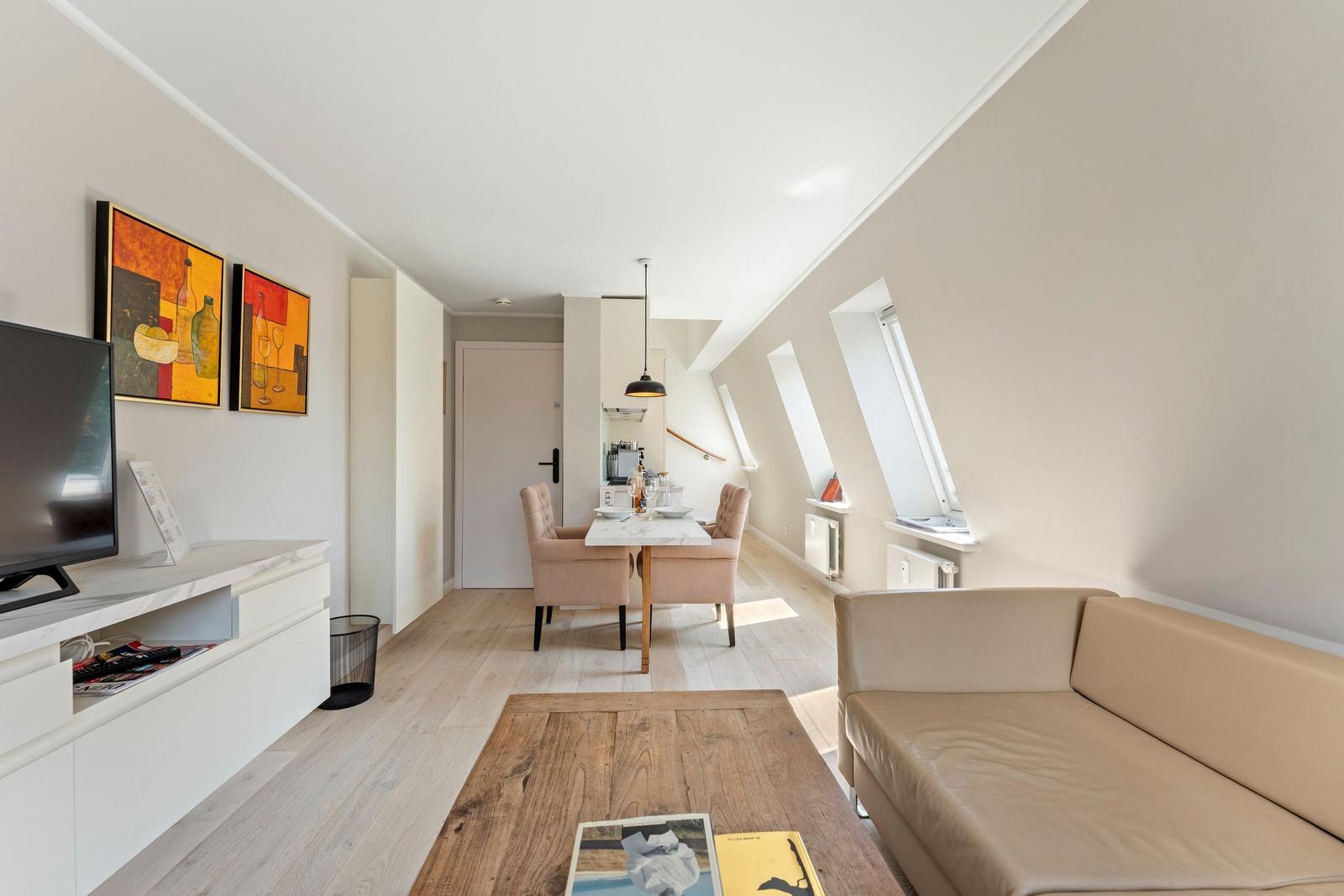 Wohn- und Essbereich mit Sofa, Tisch und Stühlen. Dachfenster und Fernseher sichtbar.