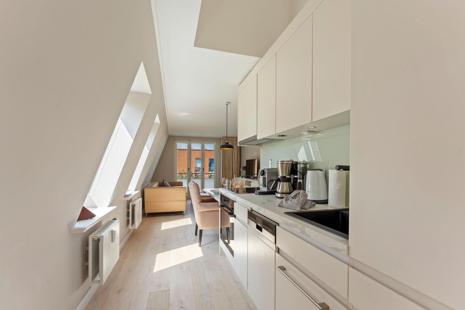 Küche mit Arbeitsfläche, Spüle und Geräten. Blick in Wohnbereich mit Sofa und Fenster.