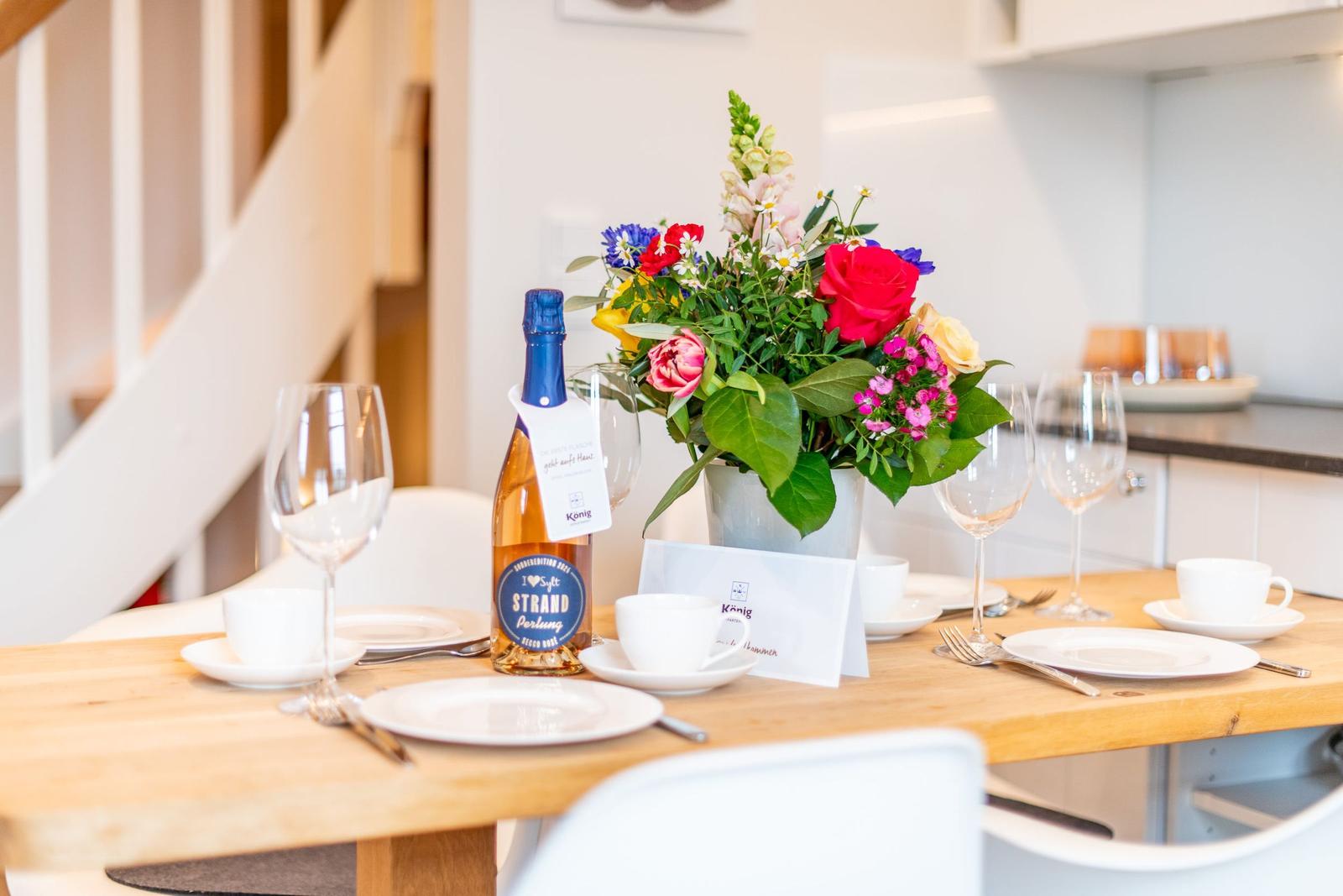 Gedeckter Tisch mit Blumenstrauß und Flasche. Hintergrund: Küchenbereich und Treppe.