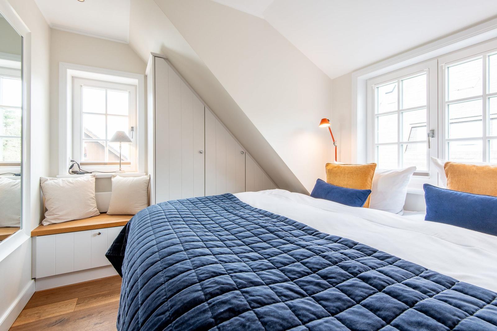 Schlafzimmer mit Dachschräge, Bett mit blauer Decke und Fenster mit Sitzbank.