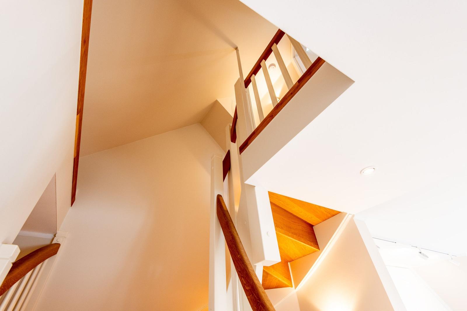 Treppe mit Holzgeländer und weißen Wänden, beleuchtet von Deckenleuchten.