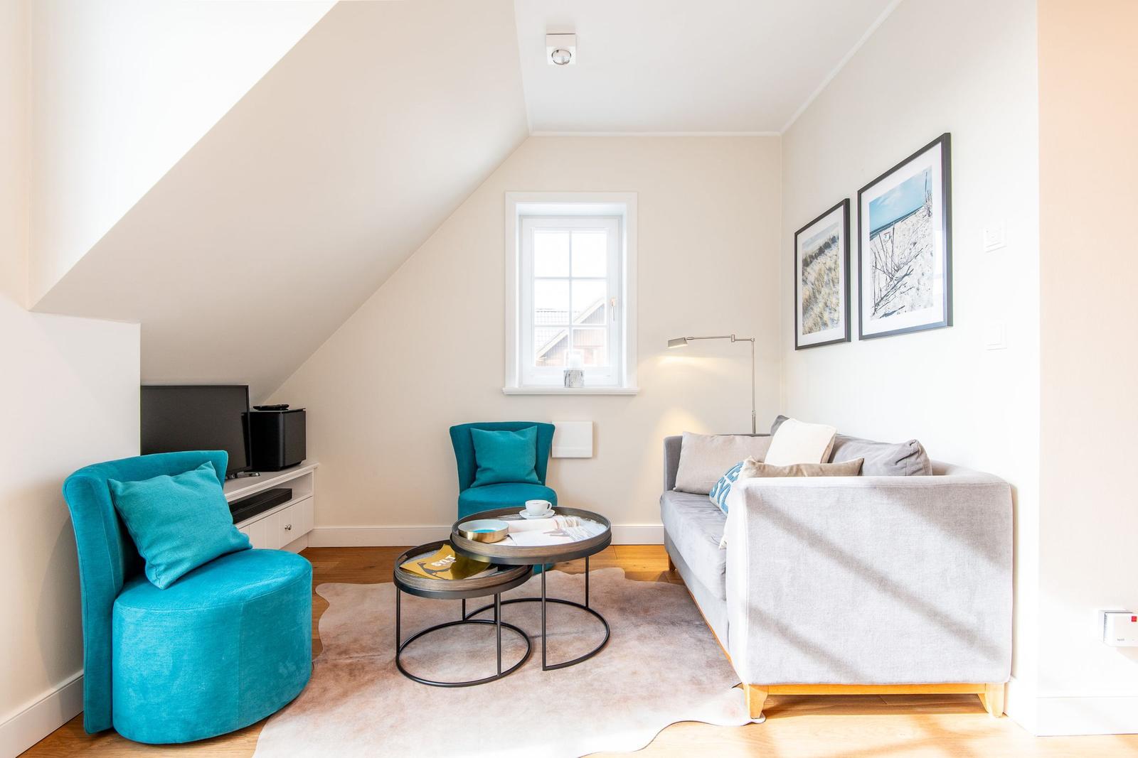 Wohnzimmer mit Sofa, zwei blauen Sesseln, Tisch und Fenster unter Dachschräge.