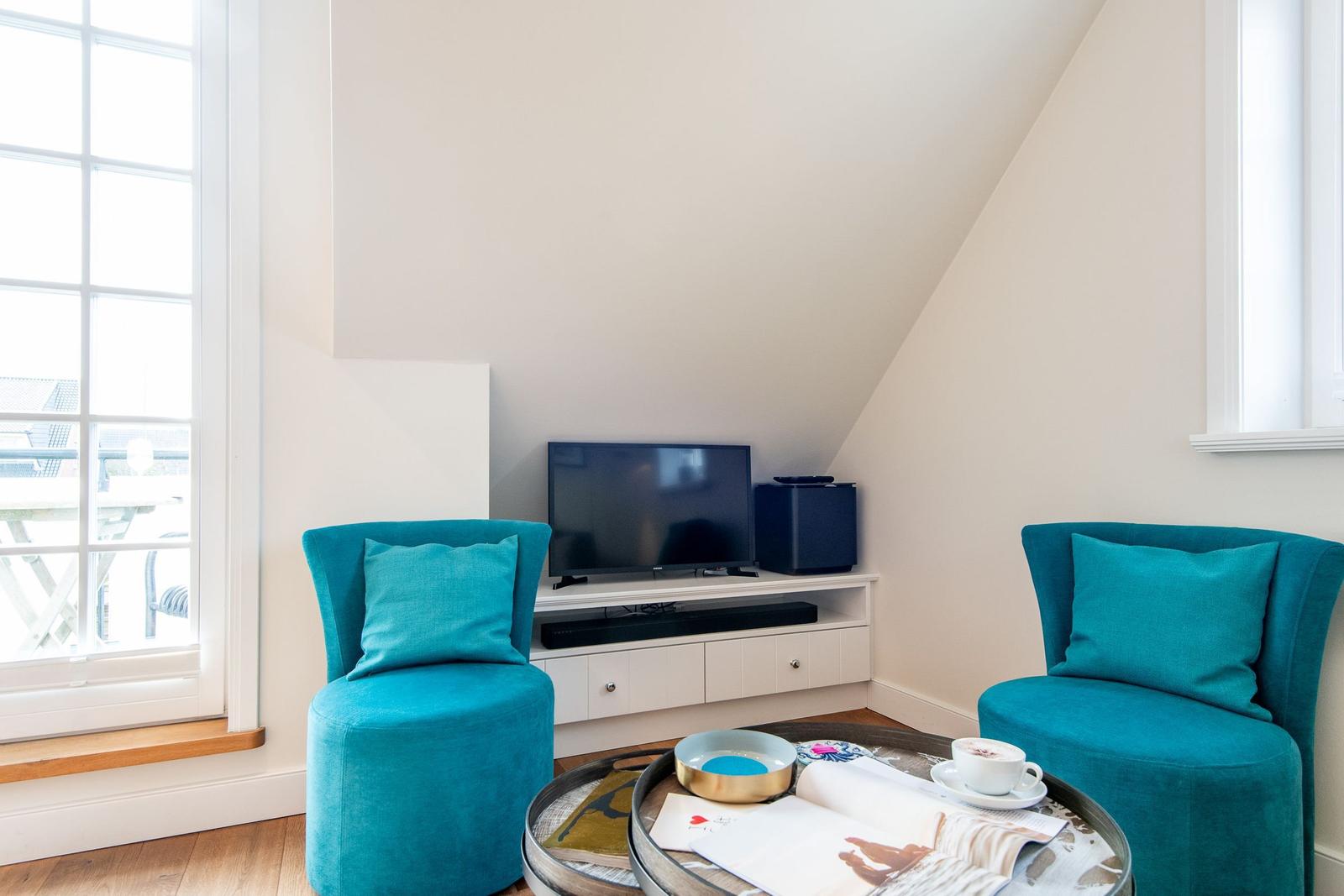 Zwei türkisfarbene Sessel stehen vor einem Fernseher. Ein runder Tisch mit Tasse und Zeitschrift.