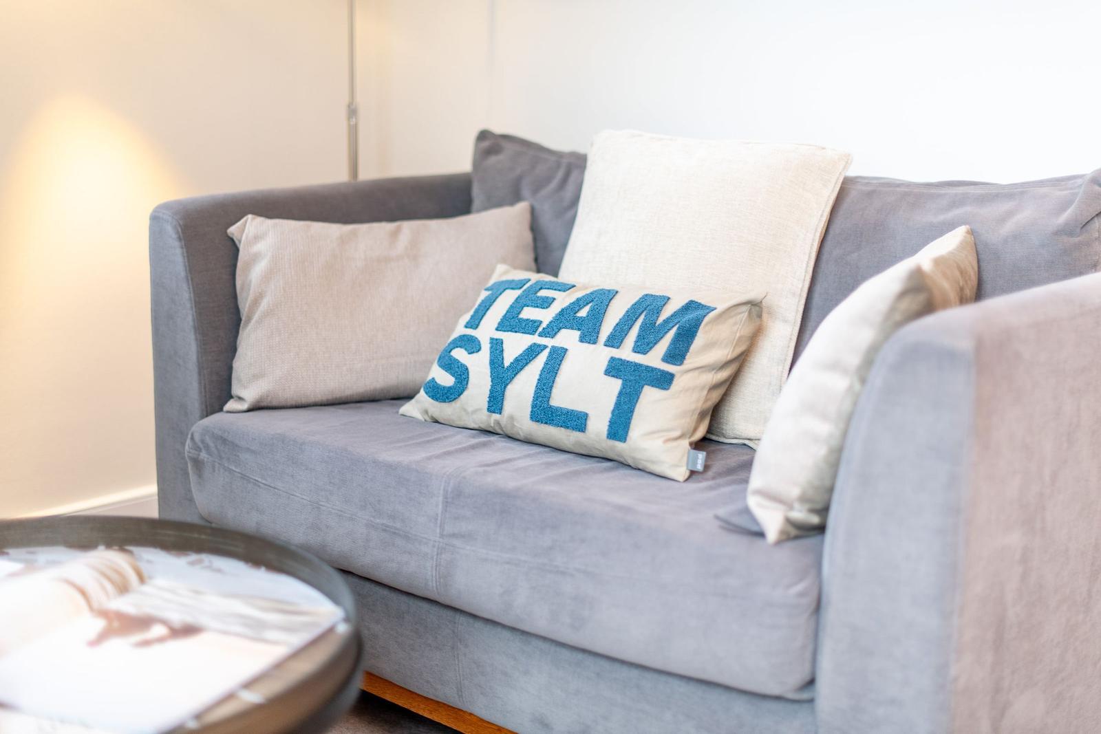 Ein grauer Sofa mit Kissen, eins mit der Aufschrift TEAM SYLT.