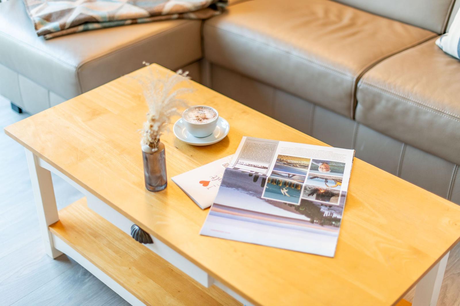 Holztisch mit Kaffeetasse, Zeitschrift und Trockenblumen vor einem Ledersofa.
