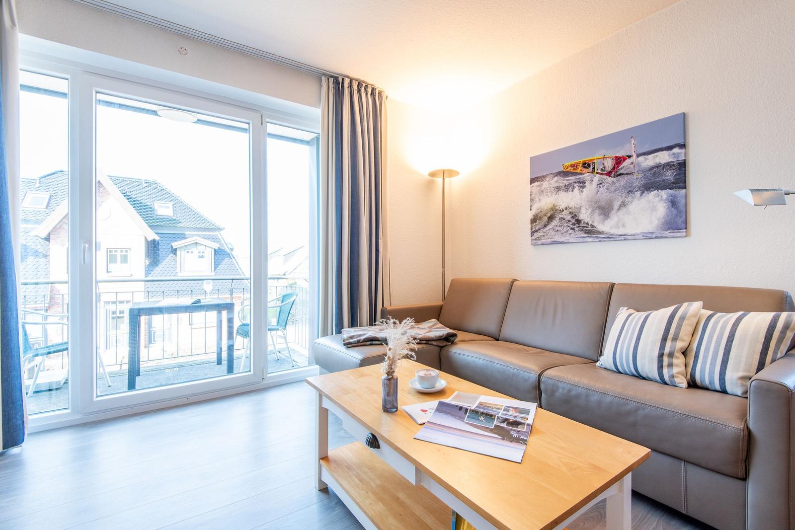 Wohnzimmer mit Sofa, Tisch und Balkonansicht. Wandbild zeigt Windsurfer.