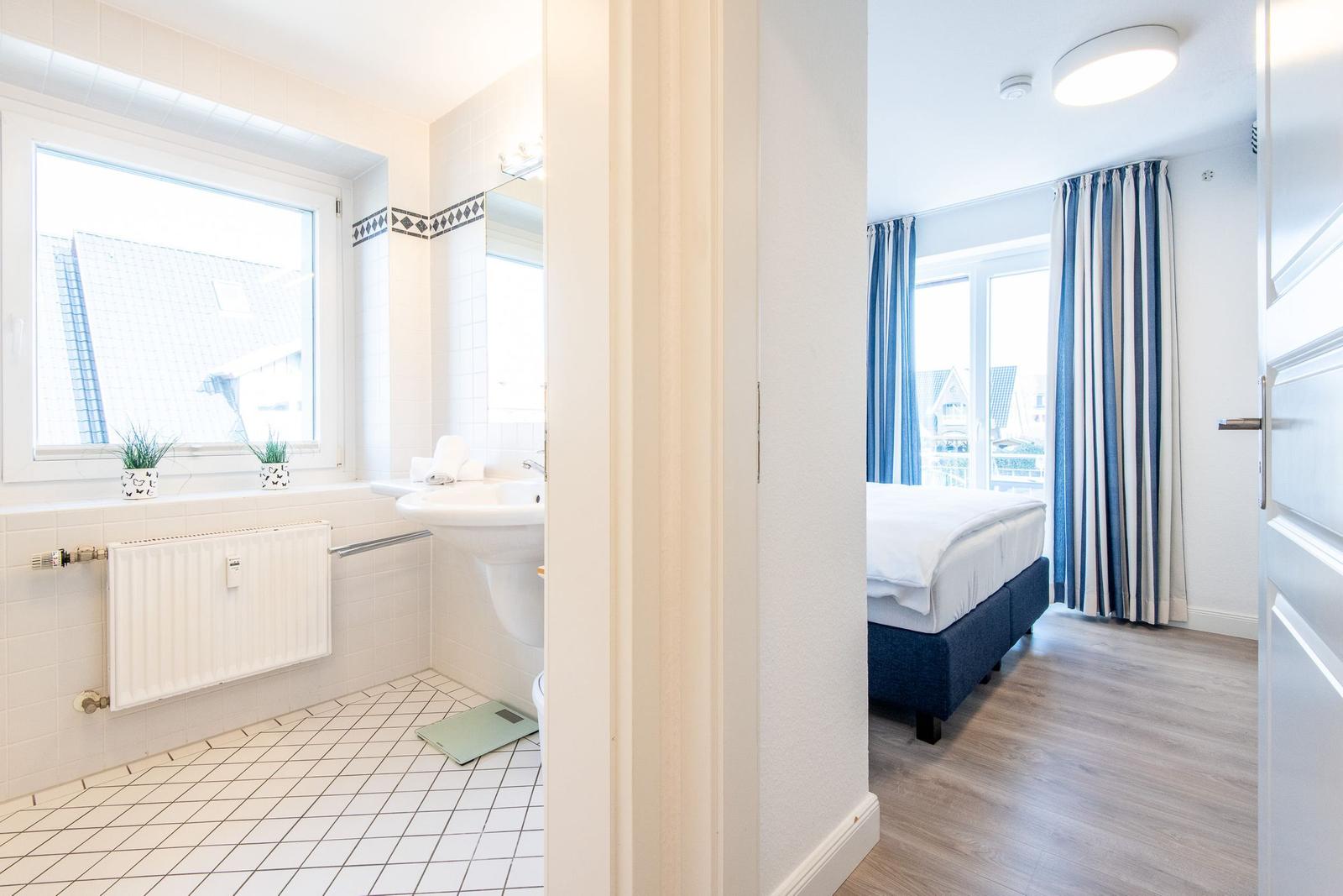 Badezimmer mit Fenster und Waschbecken. Nebenraum mit Bett und Vorhängen.