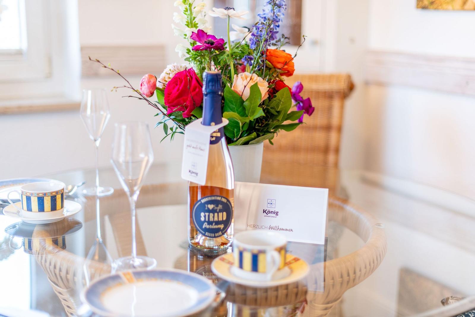 Tisch mit Blumen, Wein und Tassen auf Glasplatte.