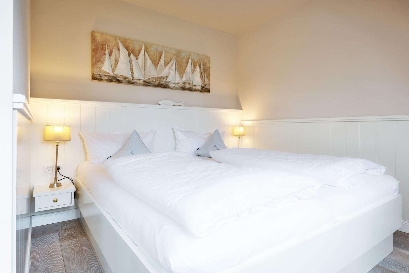 A bed with white linens, two nightstands with lamps, and a wall painting with sailboats.