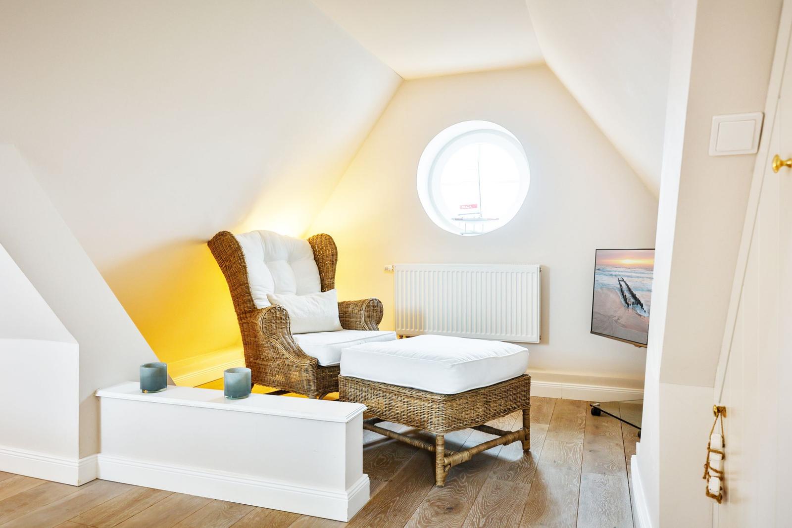 A bedroom with rattan armchair, round window and wooden floor.