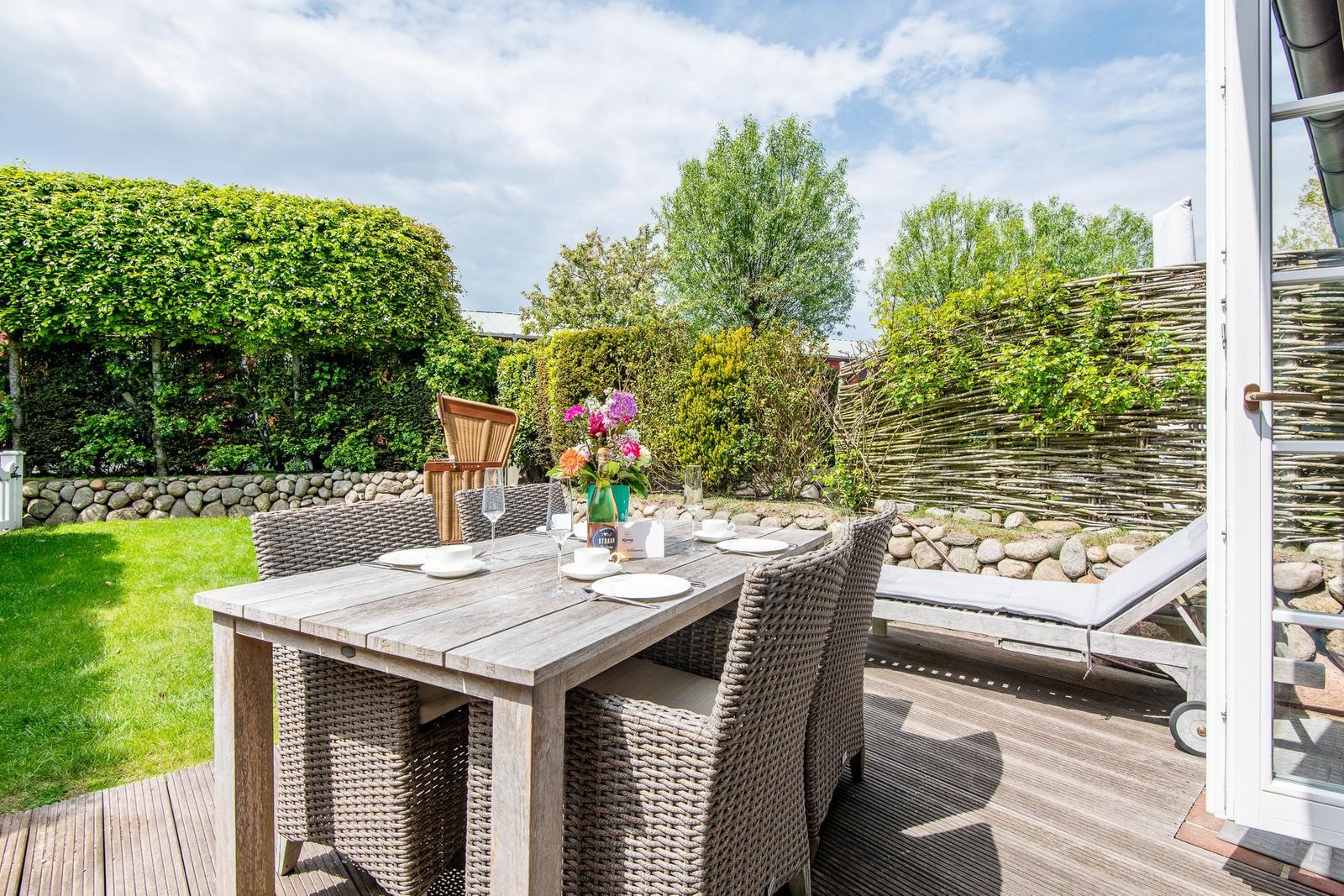 Deck with wooden table, chairs, and lounge chair. Backyard with lawn and hedges.