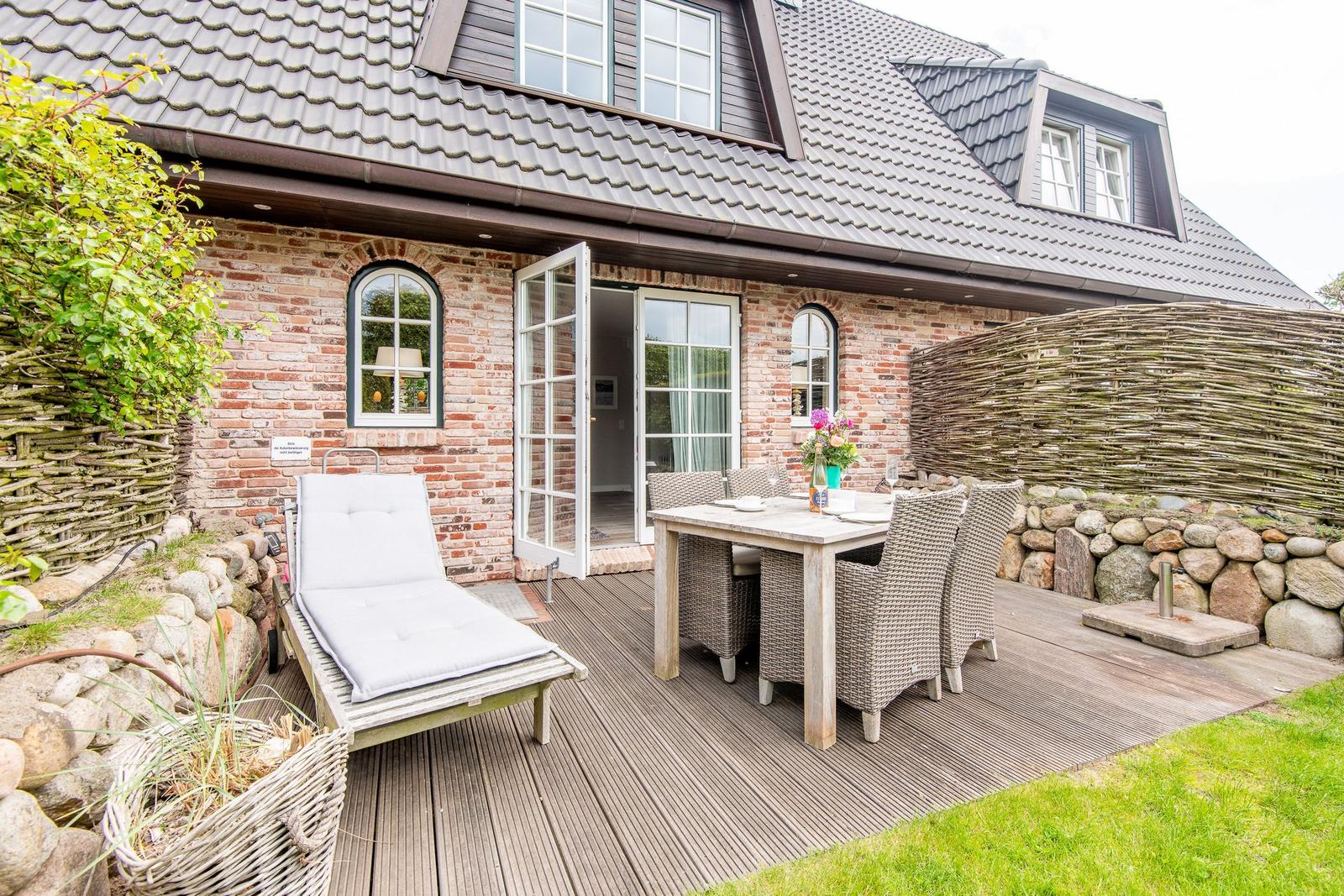 Deck with table, chairs, and lounge. House with brick wall and dormer windows.