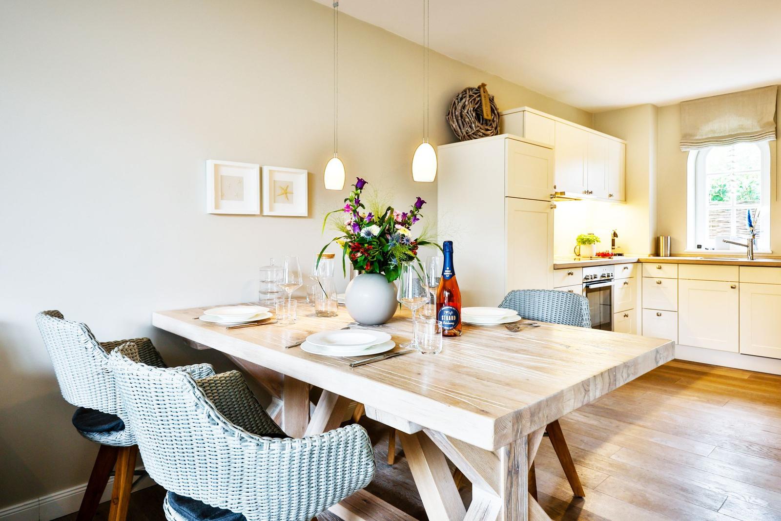 Dining area with wooden table and chairs. Kitchen in background with window.