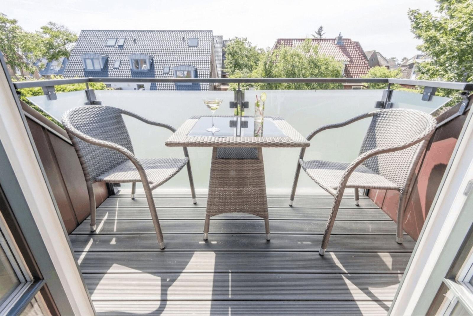 Terrasse mit Tisch und zwei Stühlen. Glasgeländer und Blick auf Wohnhäuser.