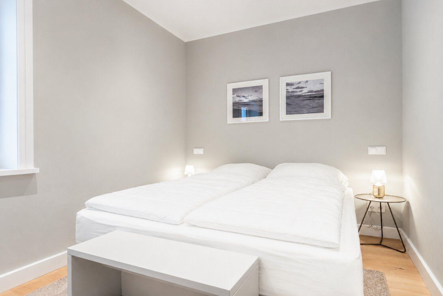Schlafzimmer mit weißem Bett, zwei Bildern an der Wand und Nachttisch mit Lampe.