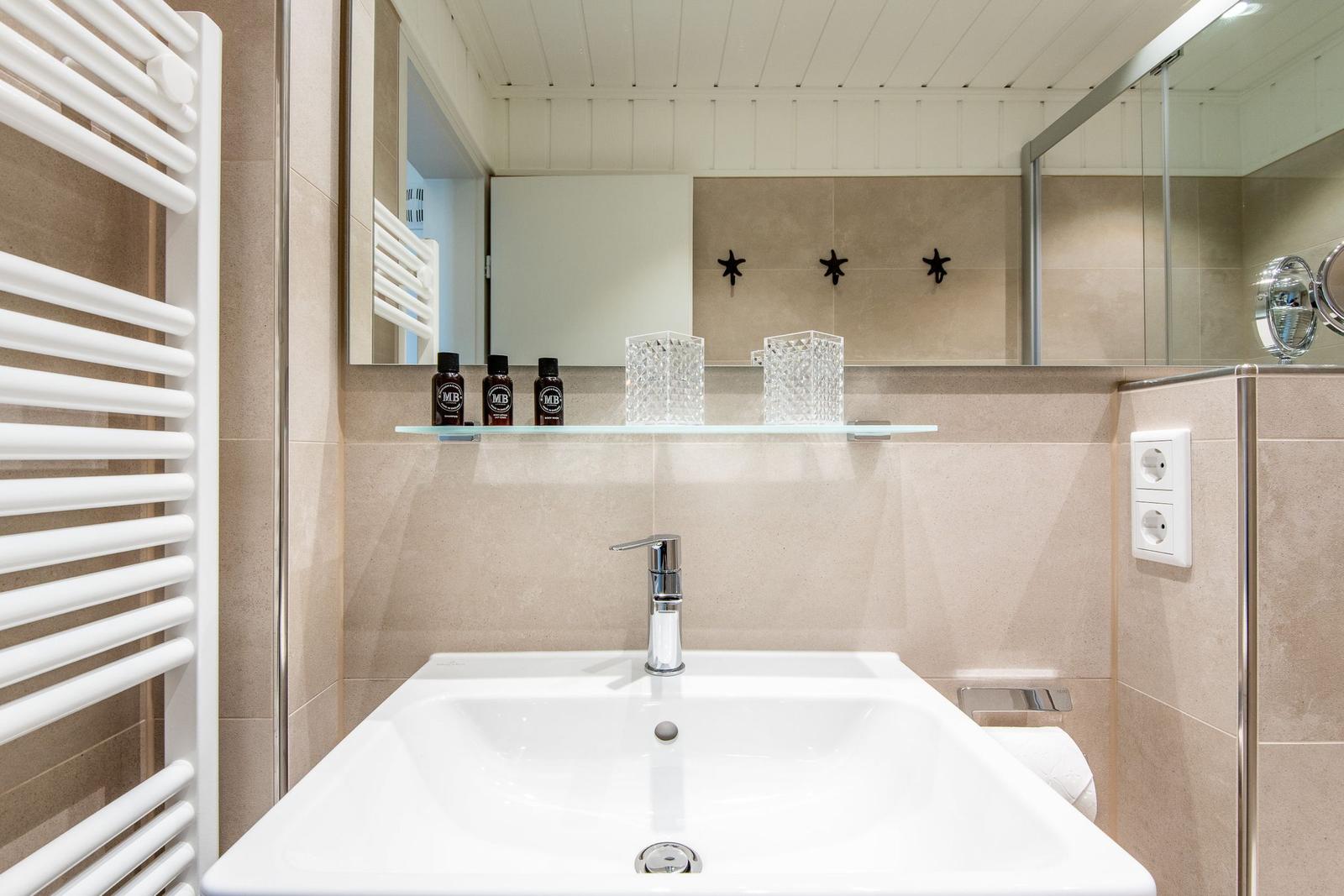 Badezimmer mit Waschbecken, Spiegel und Dusche. Drei Flaschen und zwei Vasen auf der Ablage.