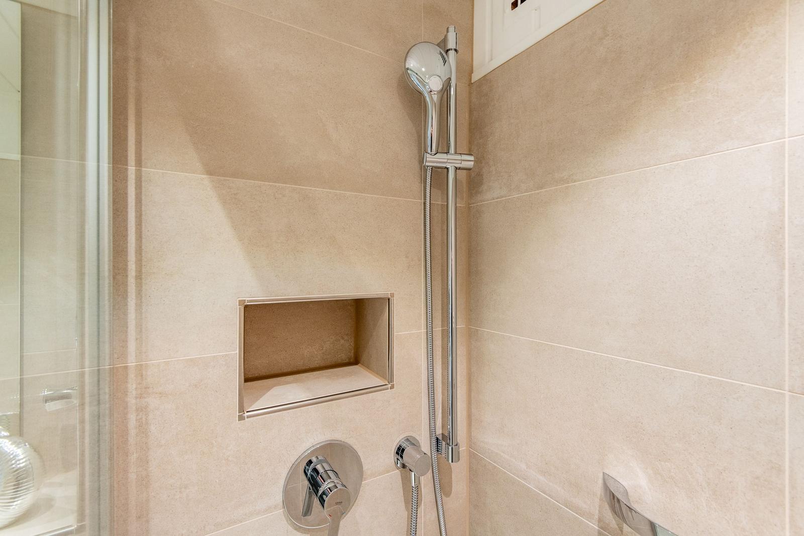 Dusche mit Handbrause und eingelassenem Regal an hellen Fliesenwänden.