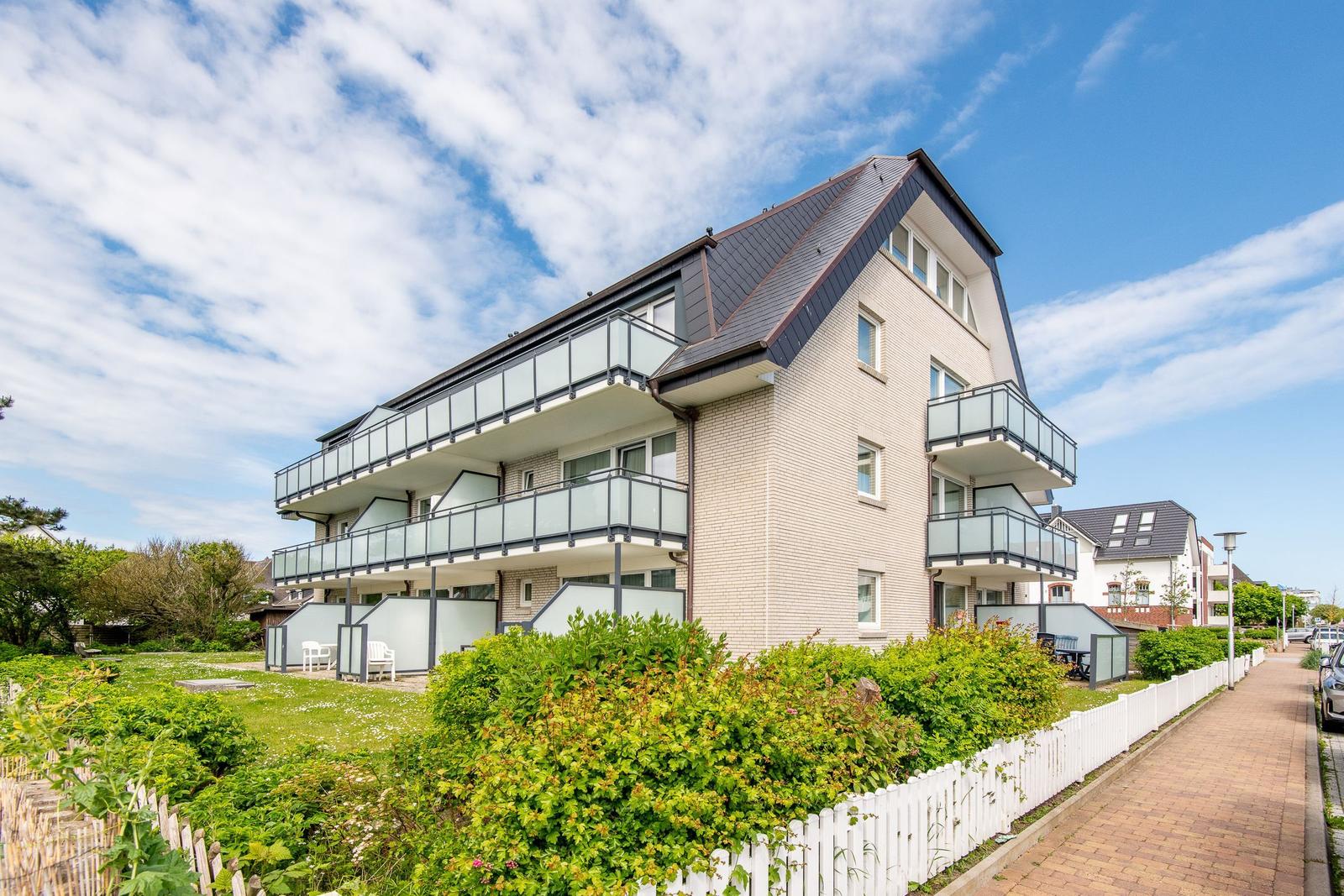 Modern multi-family house with balconies and garden area. White fences and green hedges.