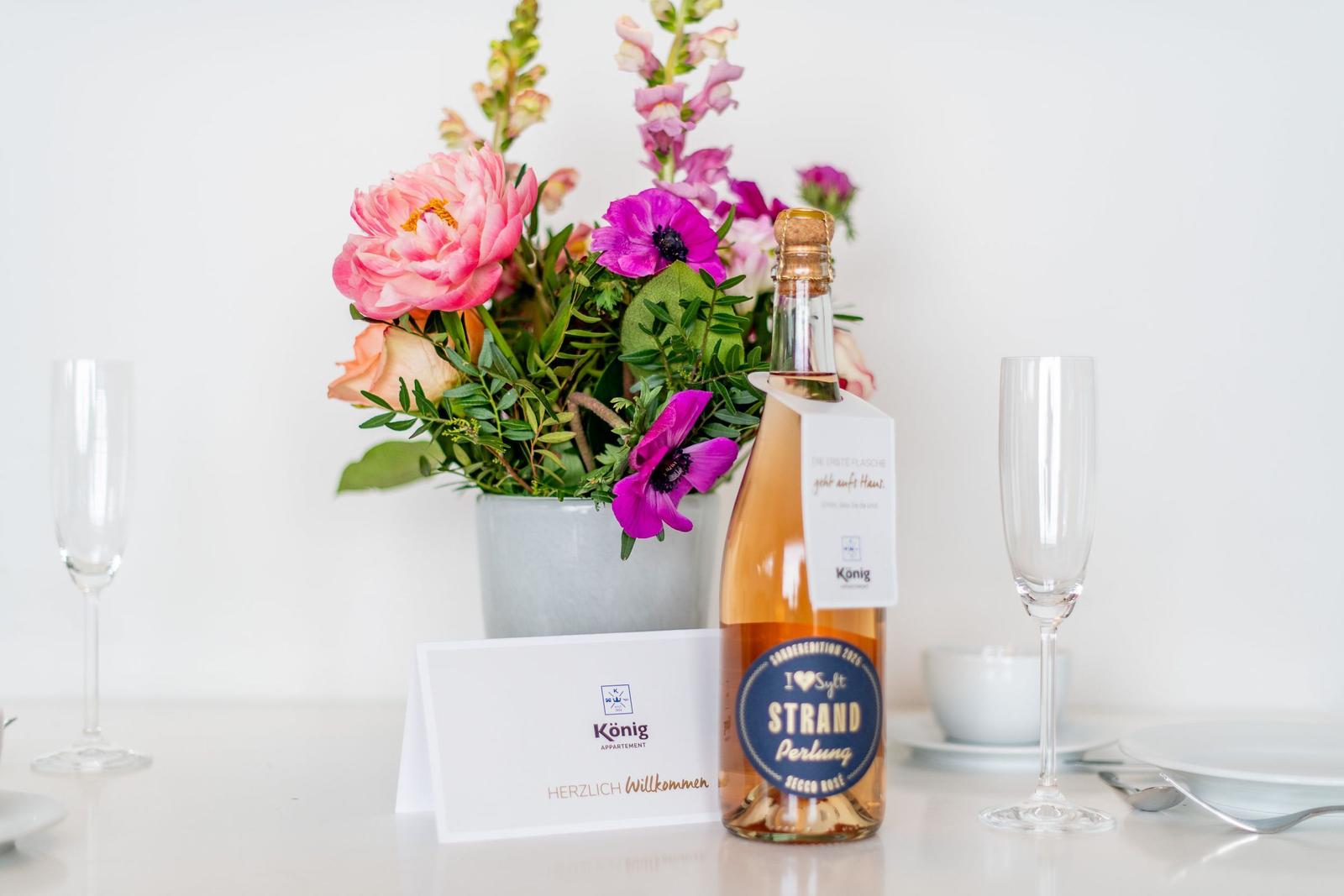 Tisch mit Blumen, Sektflasche und Gläsern. Willkommenskarte und Teller sind sichtbar.