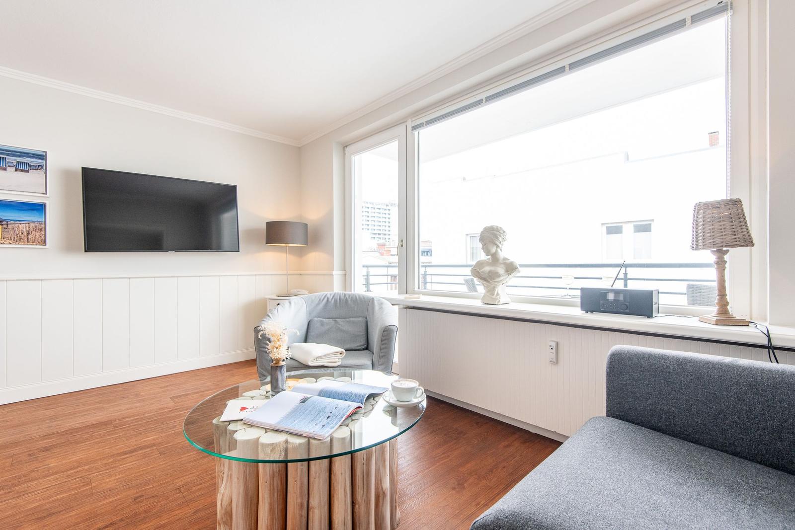 Wohnzimmer mit Fernseher, Sofa und Sessel. Glas-Tisch, Fenster und Balkon.
