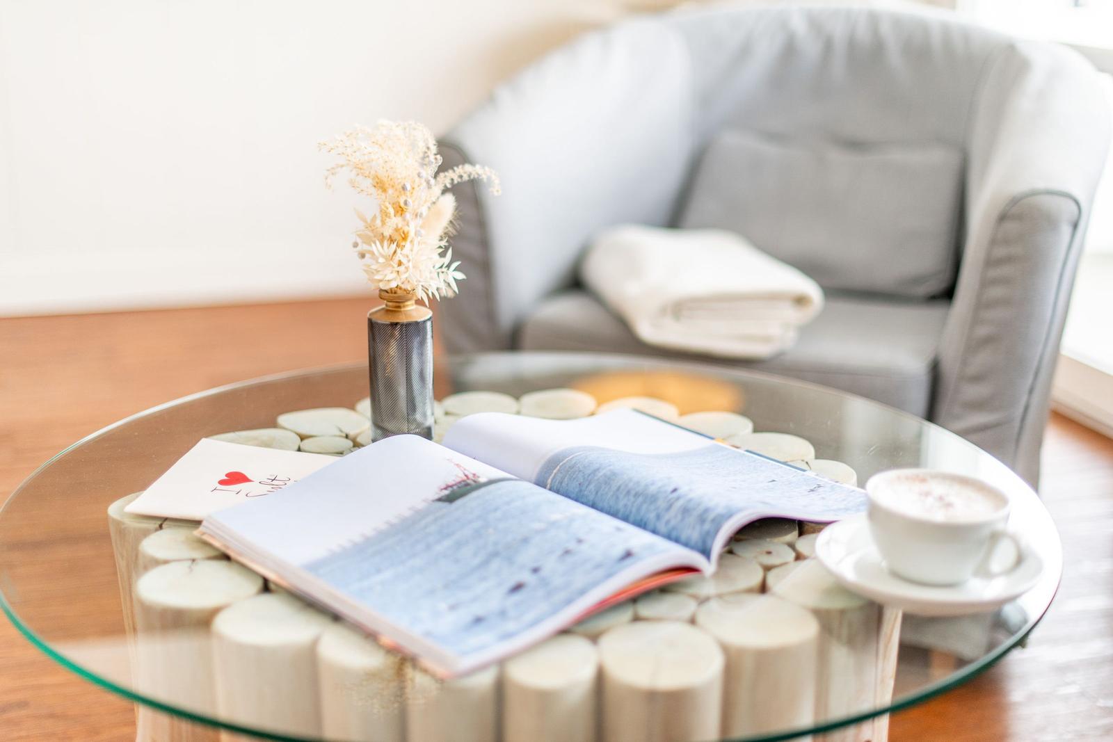 Auf dem Tisch liegt ein geöffnetes Buch, eine Tasse und eine Pflanze. Im Hintergrund ein Sessel mit einer Decke.