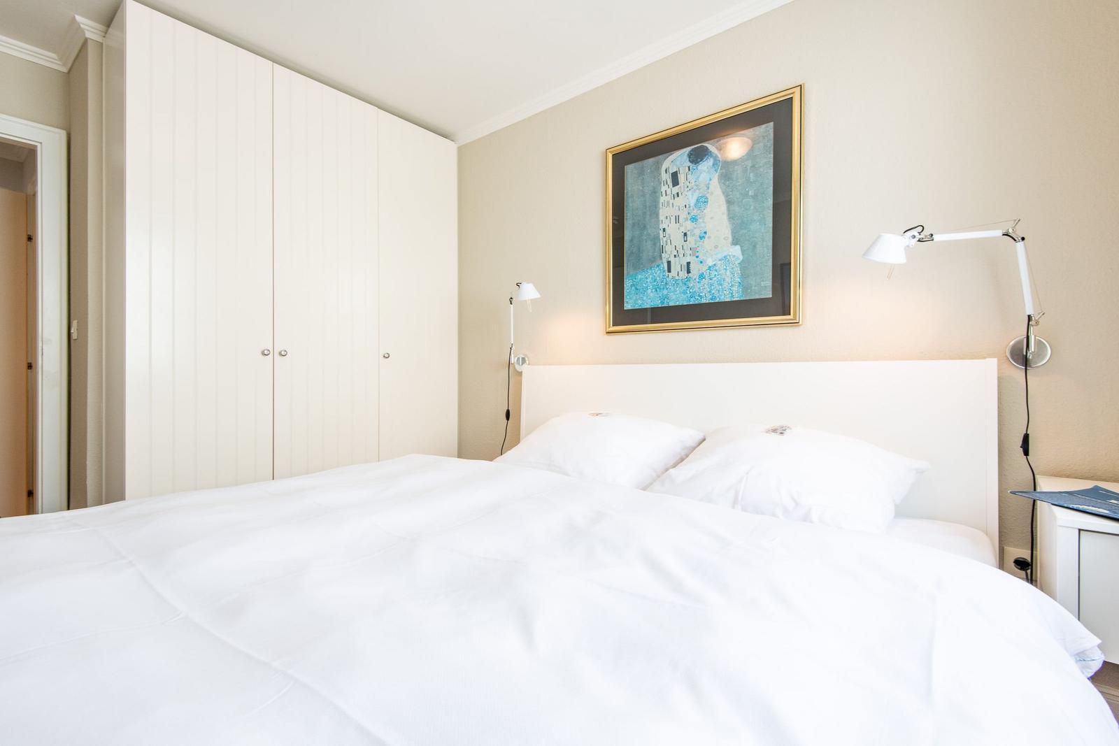 Schlafzimmer mit weißem Bett, Kleiderschrank und Wandbild.