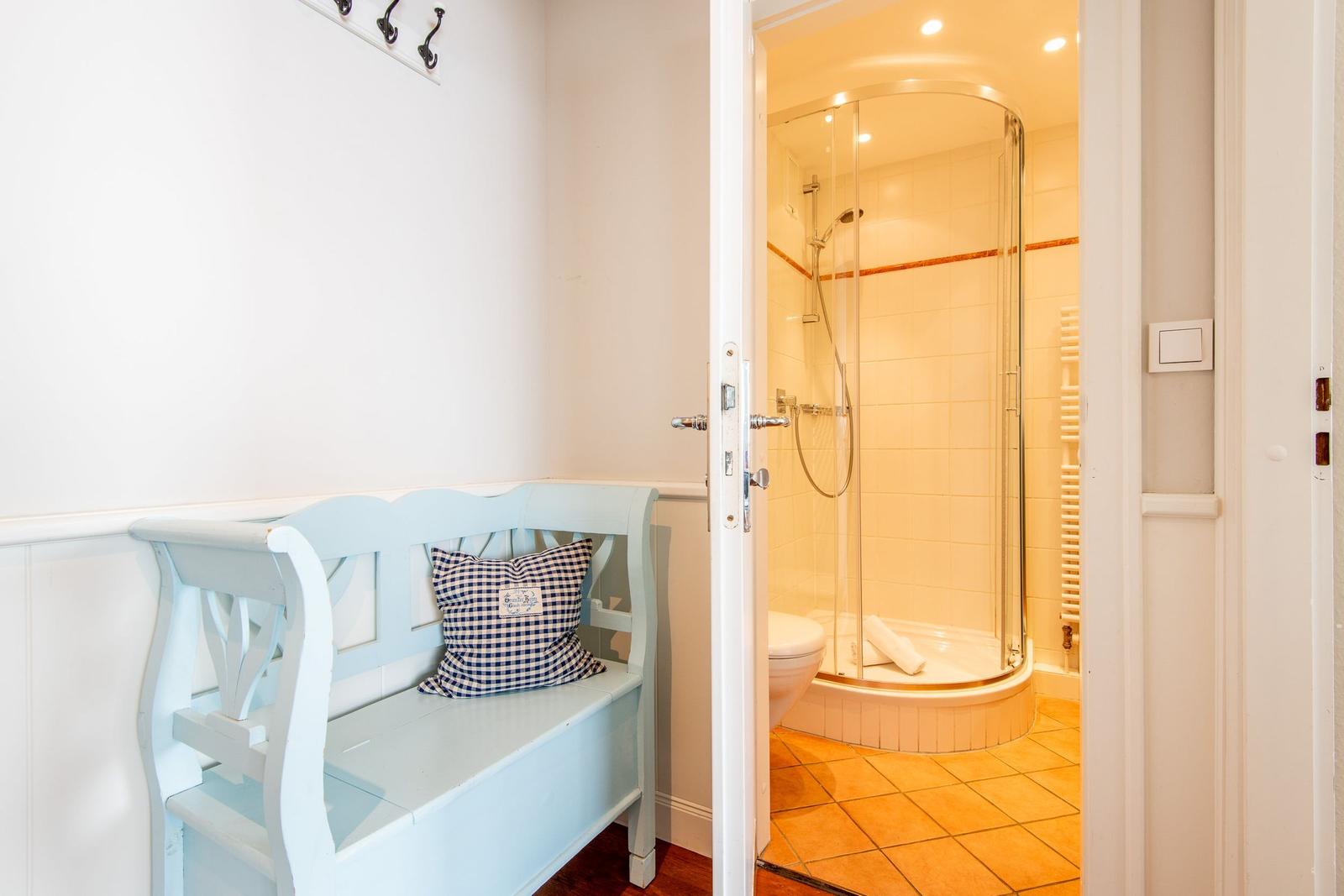 Helle Dusche mit Glasabtrennung und Toilette. Vorne eine blaue Bank mit Kissen.