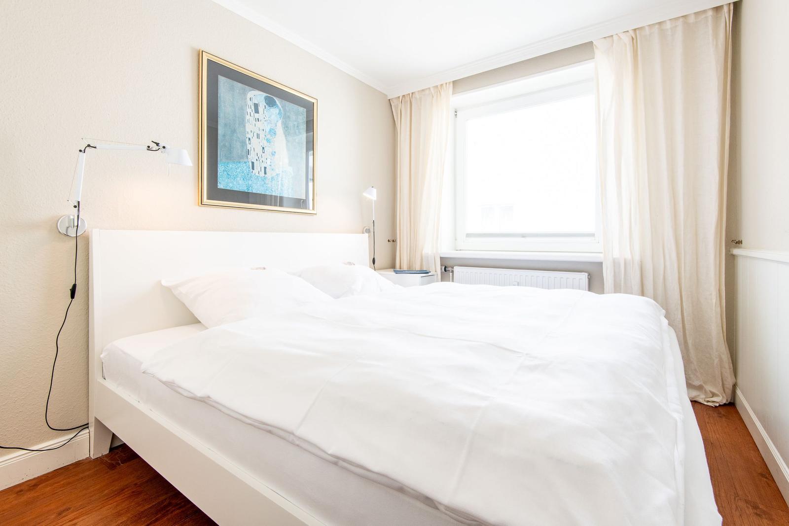 Schlafzimmer mit weißem Bett, Fenster mit Vorhängen und Wandbild.