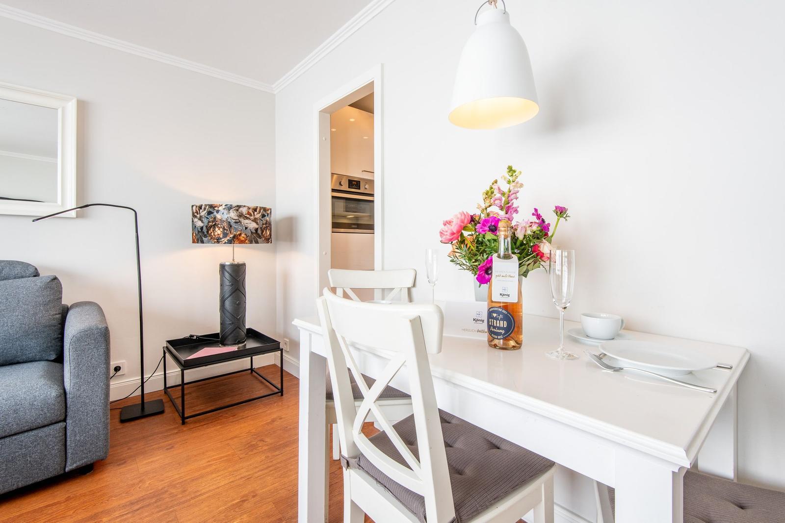 Essbereich mit weißem Tisch und Stühlen, Blumen und einer Lampe. Küche im Hintergrund.